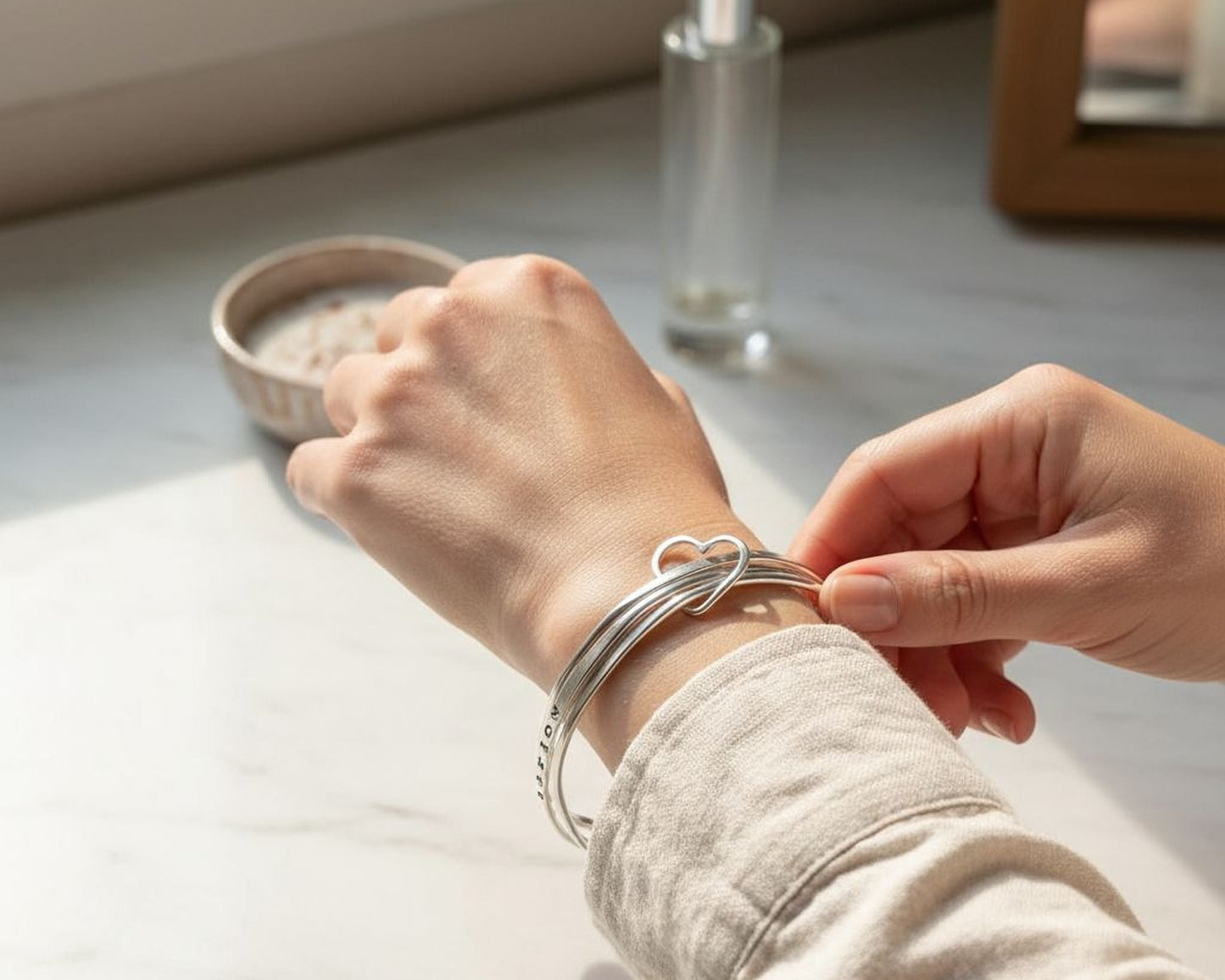 A person's hand is adjusting a silver bracelet on their wrist, with a small bowl and a bottle of perfume visible in the background.
