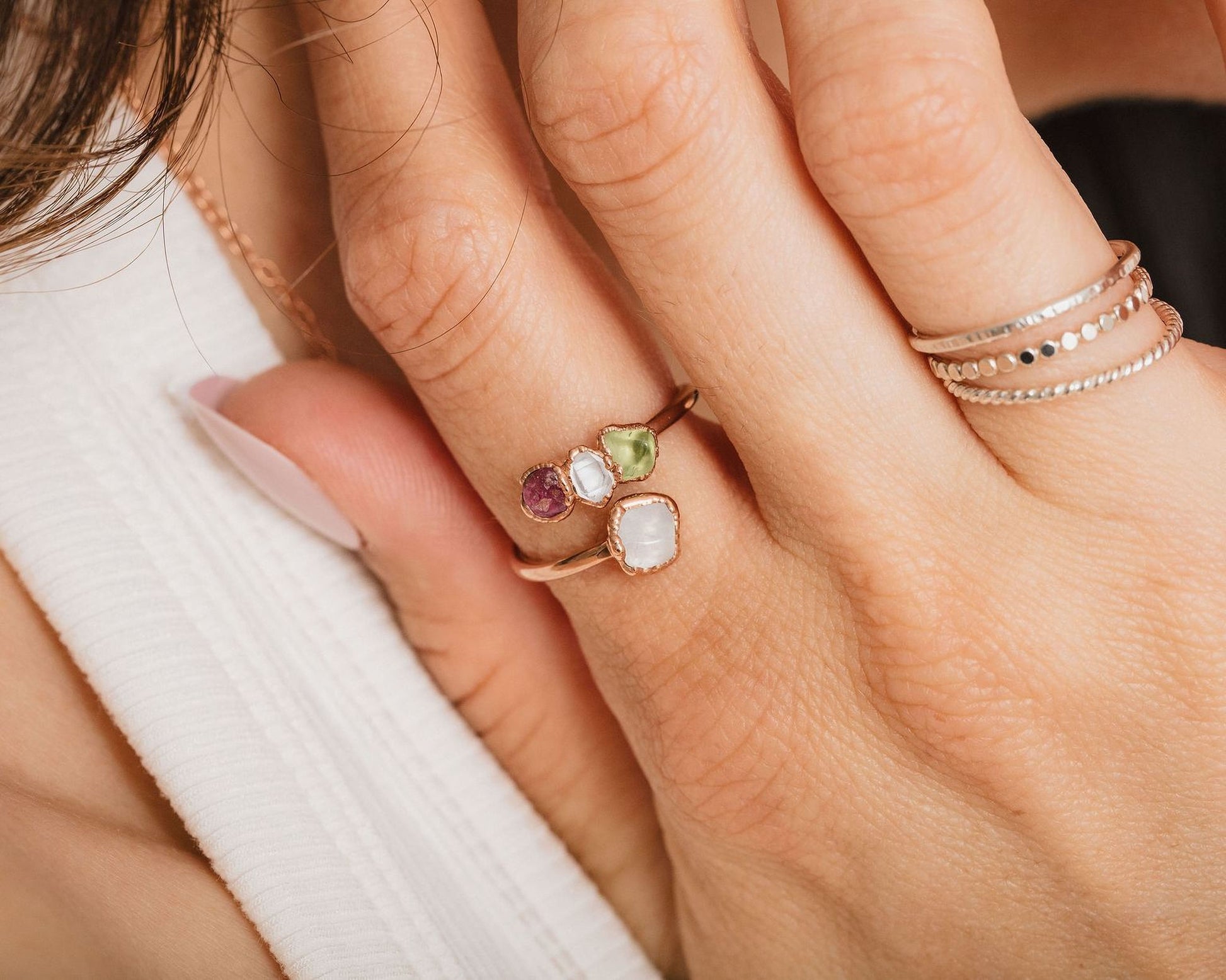A close-up of a person's hand wearing a ring with three gemstones, including a white stone, a purple stone, and a green stone.