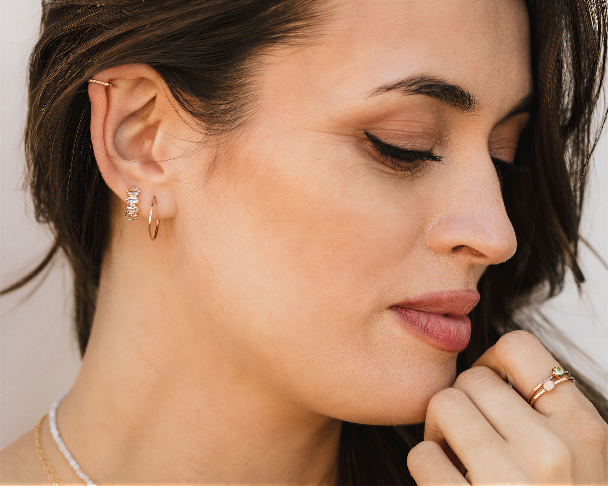 A close-up of a woman's face, with her eyes closed and her hand resting on her chin. She is wearing multiple rings on her fingers, including a large hoop earring and a ring on her ring finger.