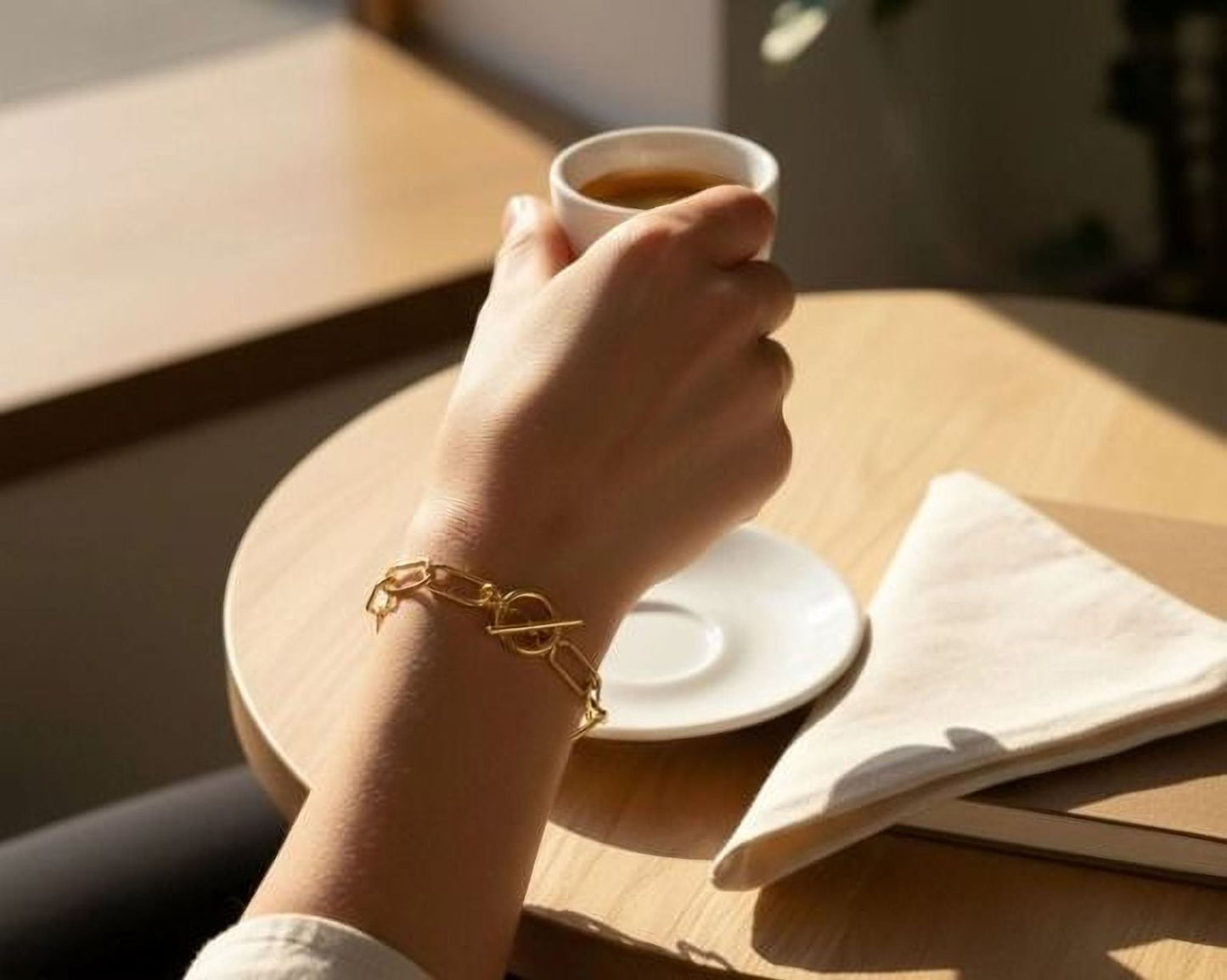 A person's hand wearing a gold bracelet is holding a cup of coffee on a wooden table, with a white plate and napkin nearby.