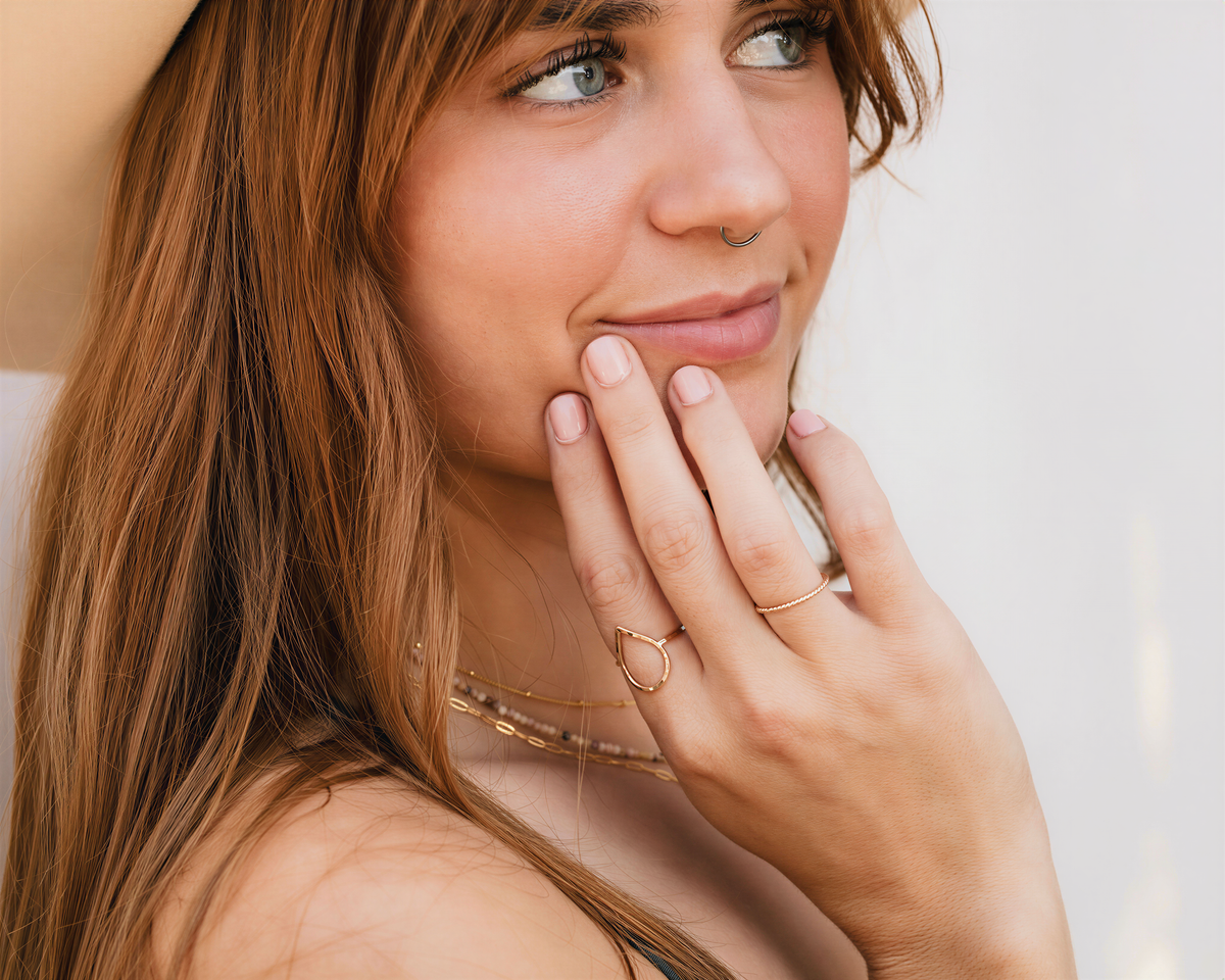 A close-up of a woman's face, with her hand resting on her chin, wearing multiple rings and a necklace.
