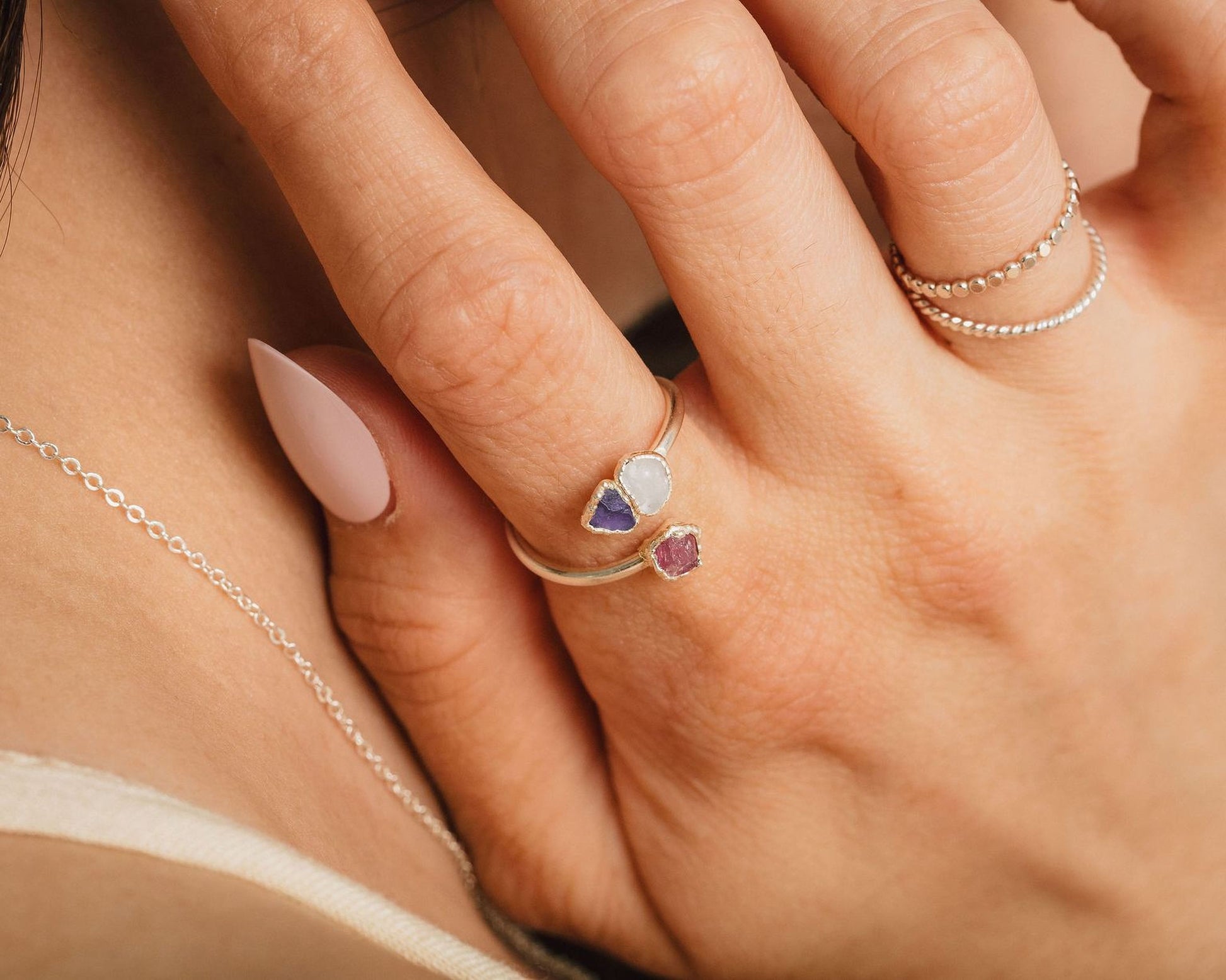 A close-up of a person's hand wearing a ring with two gemstones, one purple and one pink, and a necklace around the neck.