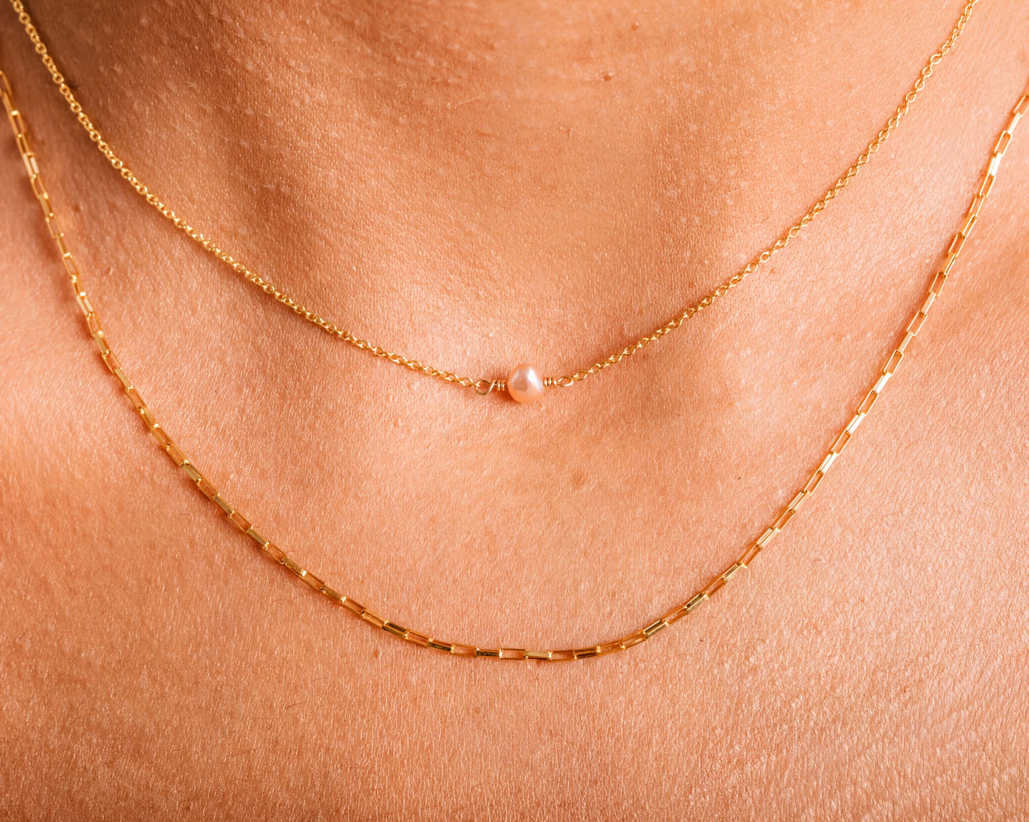 A close-up view of a person's neck adorned with two gold necklaces. The necklace on the left is a delicate chain with a single pearl pendant, while the necklace on the right is a longer chain with multiple pearls.