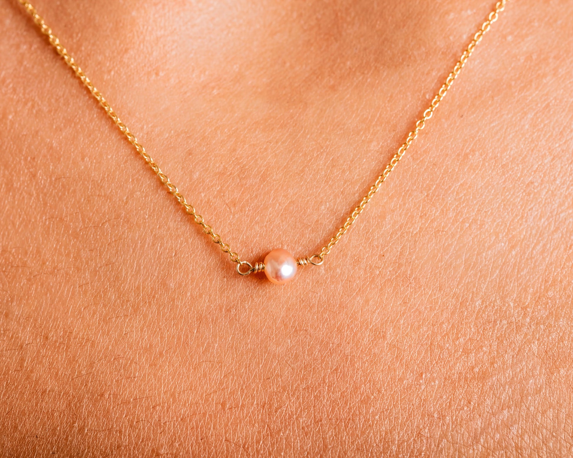 A delicate gold necklace with a single pearl pendant is shown against a textured skin background.