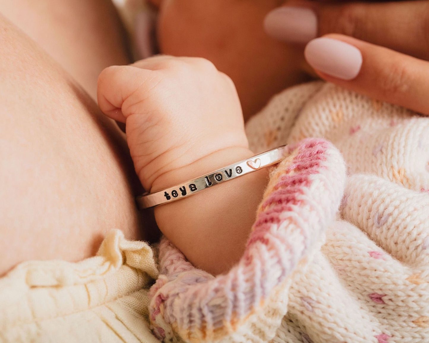 A baby's hand wearing a bracelet with the word "love" engraved on it, resting on a soft, knitted blanket.