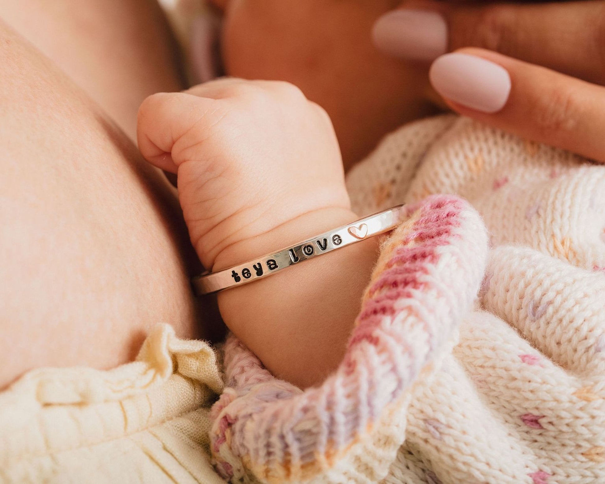 A baby's hand wearing a bracelet with the word "love" engraved on it, resting on a soft, knitted blanket.