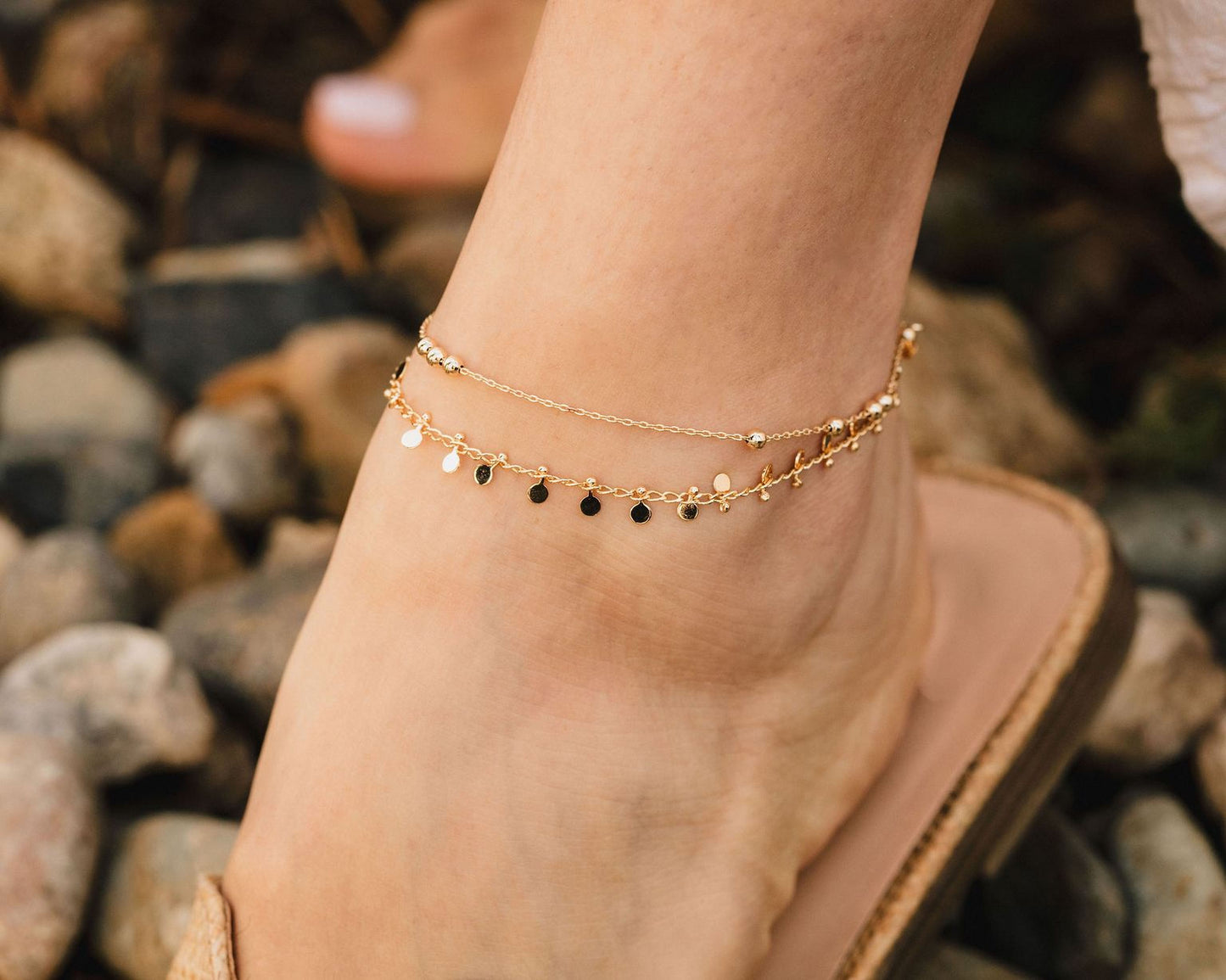 A close-up of a person's ankle wearing a gold ankle bracelet with multiple dangling charms.