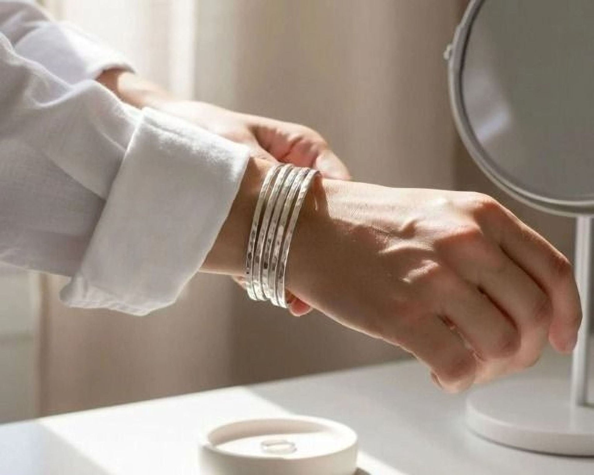 A person's hand wearing multiple silver bracelets is reaching for a white bowl on a white surface, with a round mirror in the background.