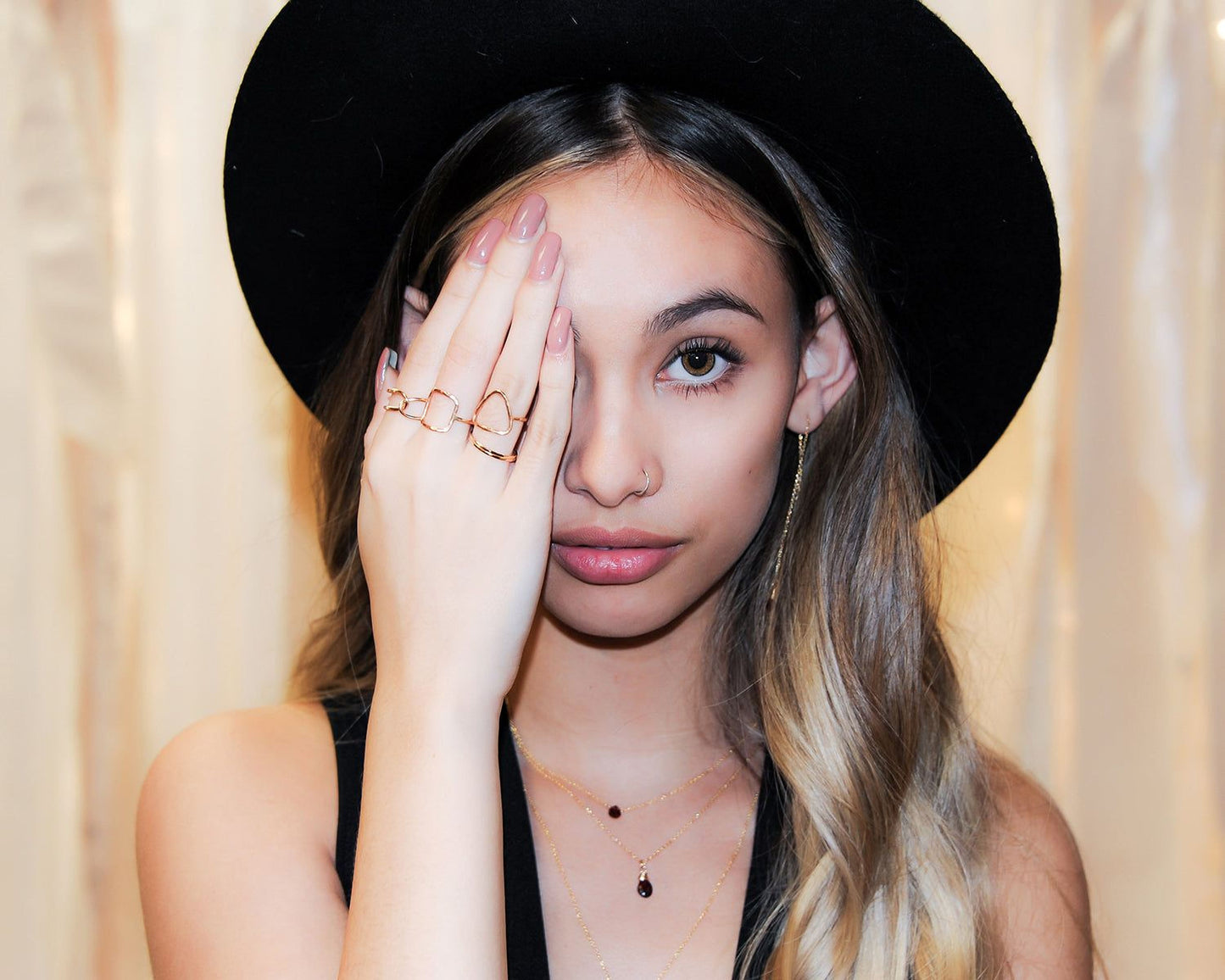 A young woman with long blonde hair wearing a black hat and multiple rings on her fingers is partially covering her face with her hand.