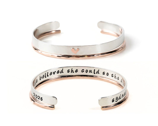 two silver-colored bracelets with a heart-shaped charm on the top. The text on the bracelets reads "She believed she could so she tried and failed and tried again."