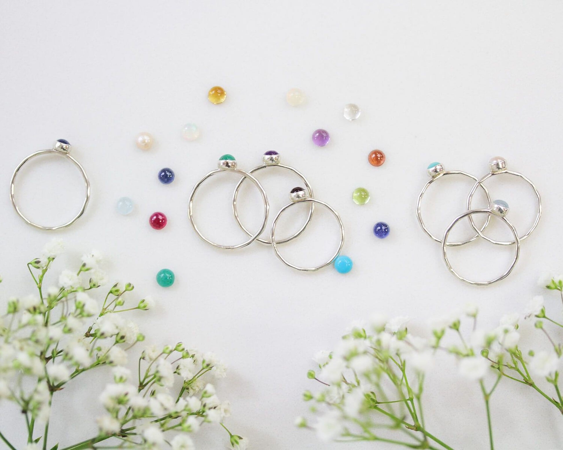 a collection of rings and gemstones, including silver rings with gemstones, scattered on a white background alongside delicate white flowers.