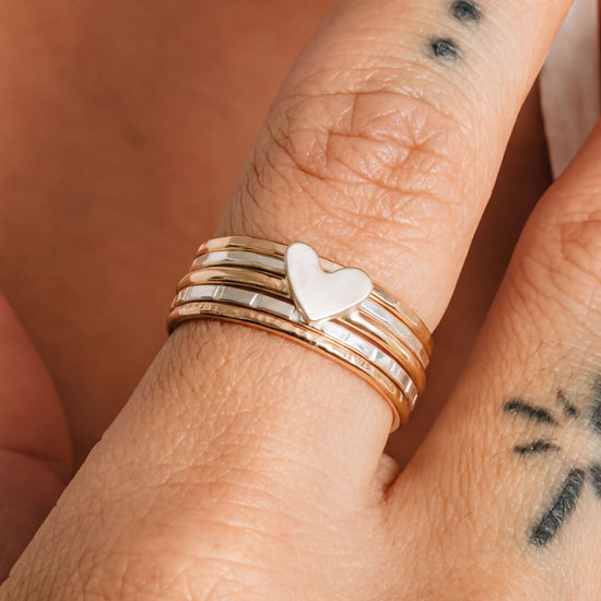 A close-up of a person's hand wearing multiple rings, including a ring with a heart-shaped charm and another ring with a tattoo design.