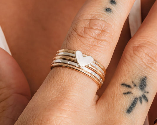 A close-up of a person's hand wearing multiple rings, including a ring with a heart-shaped charm and another ring with a tattoo design.