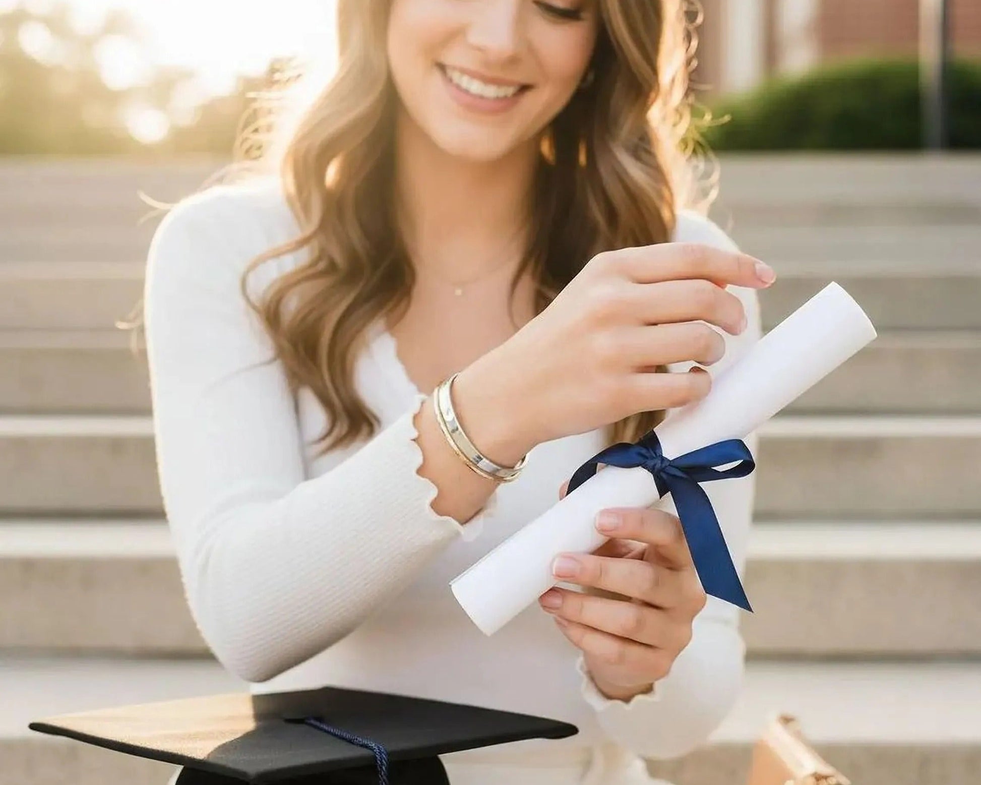 A smiling young woman holding a diploma and wearing a graduation cap sits on a set of stairs, with a brown purse nearby.