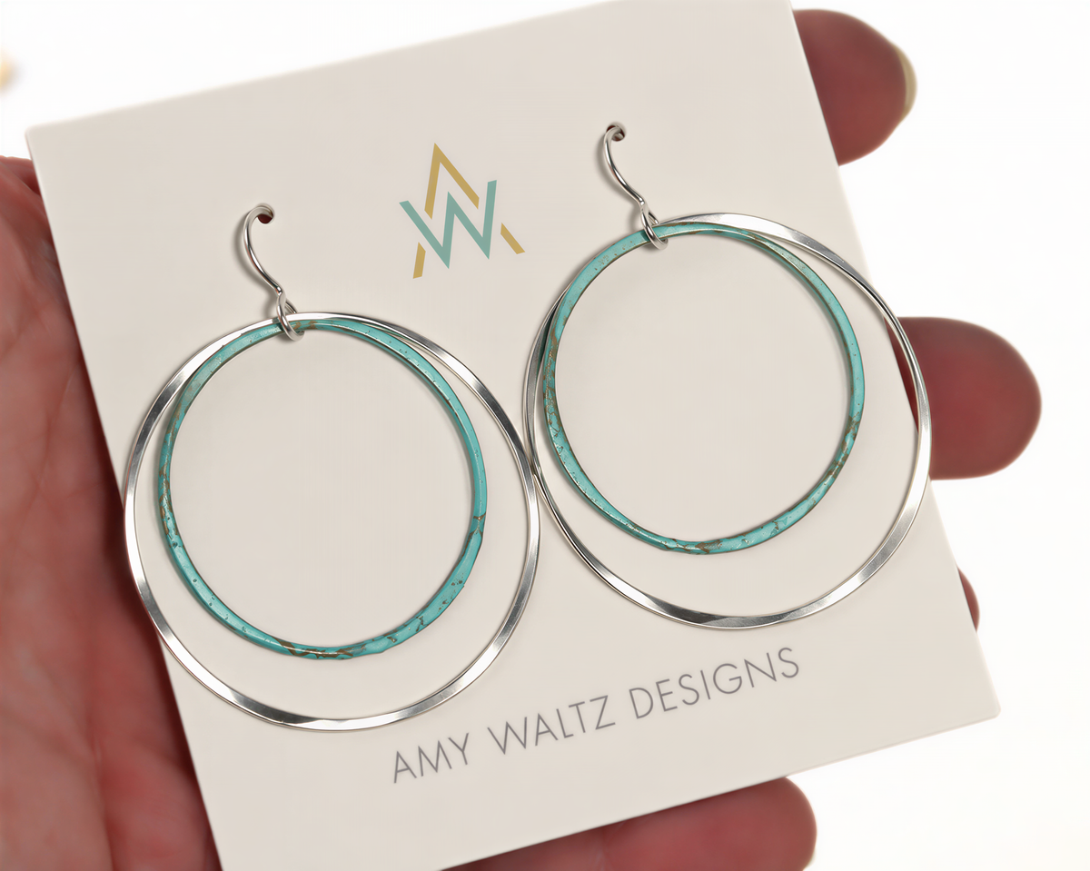 A pair of turquoise and silver hoop earrings being held in a hand, with a white card displaying the earrings and the company name.