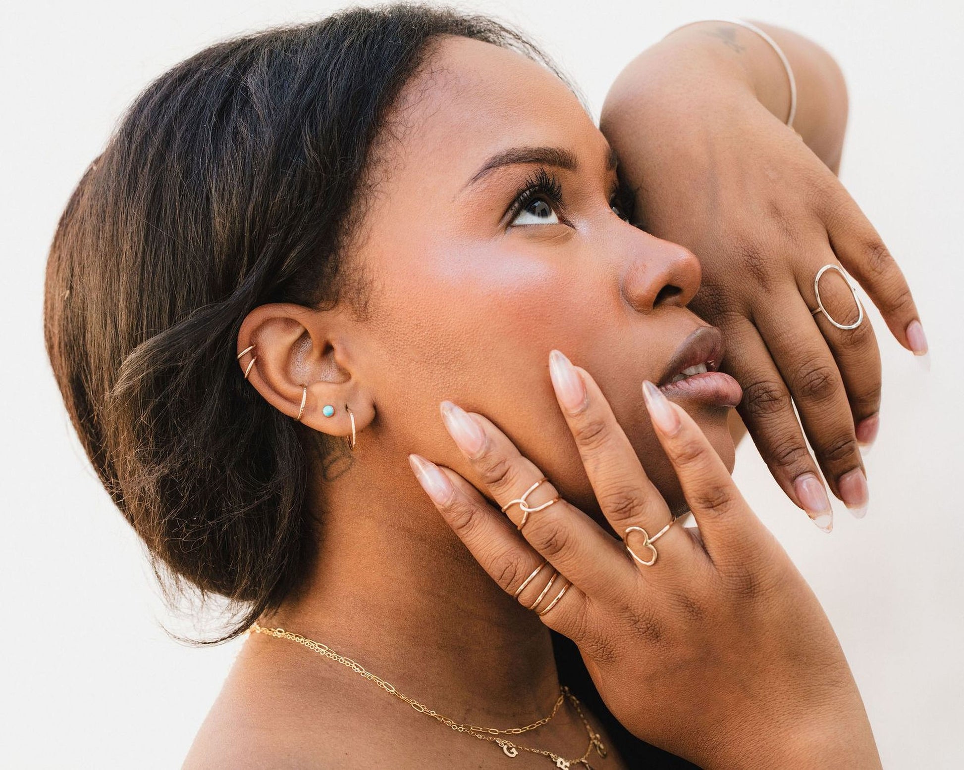 A close-up portrait of a woman with dark hair, wearing multiple rings on her fingers and a necklace around her neck.