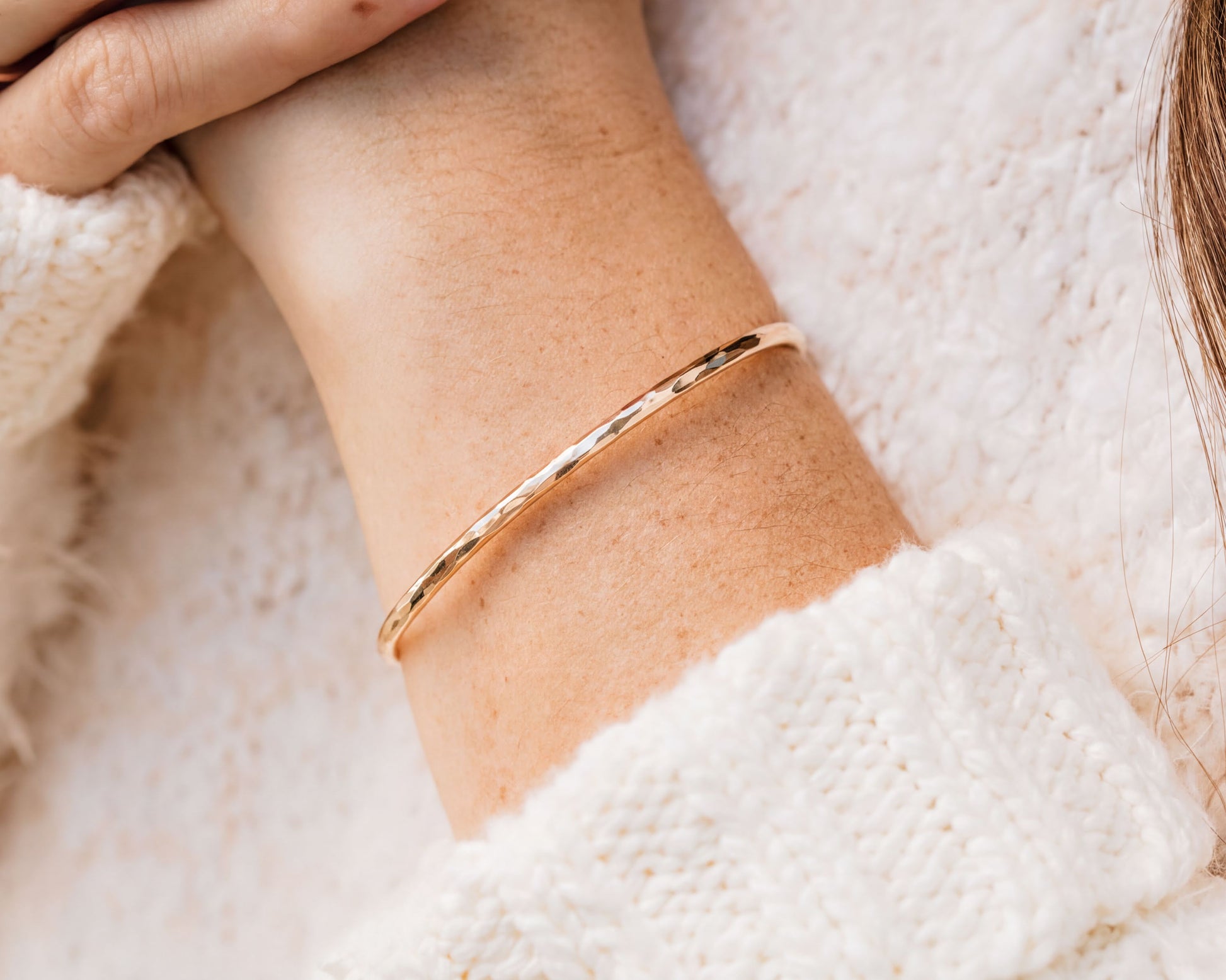 A gold-colored bracelet is worn on a person's wrist, with a white sweater visible in the background.