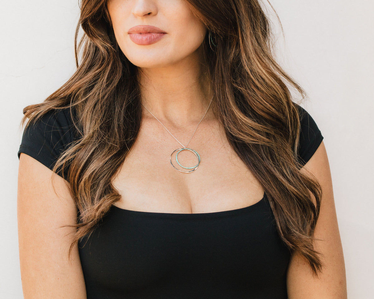 A woman with long, wavy brown hair wearing a black top and a necklace with a circular pendant.