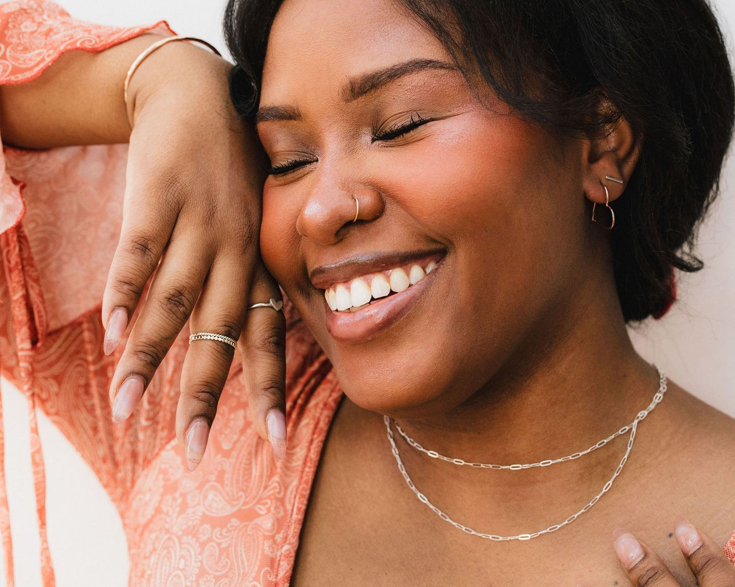 A woman with a bright smile and dark hair is wearing a pink patterned blouse and multiple necklaces, with her eyes closed and her hand resting on her face.
