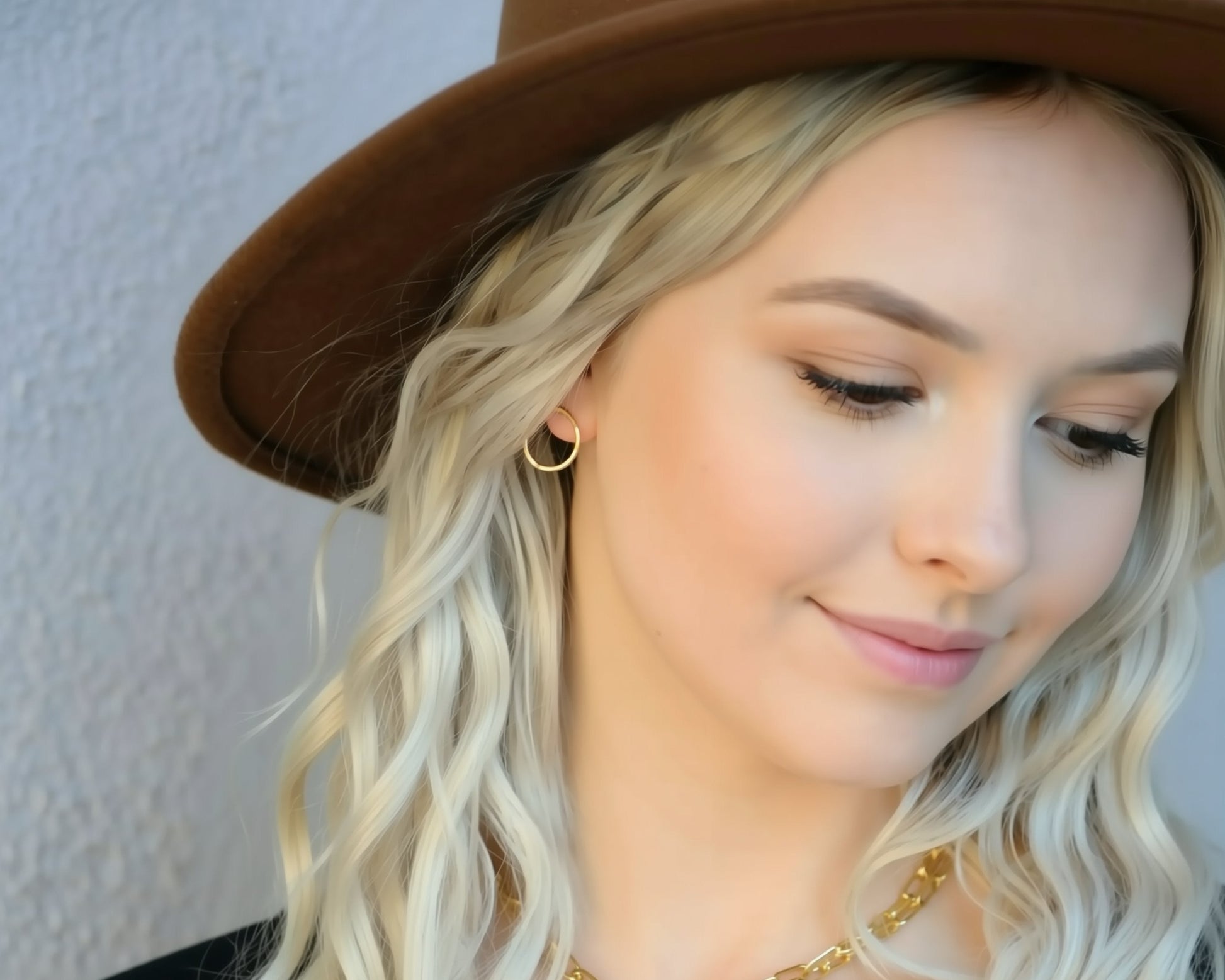 A close-up of a woman's face, with her blonde hair and a brown hat. She is wearing a gold-colored earring and a gold-colored necklace.