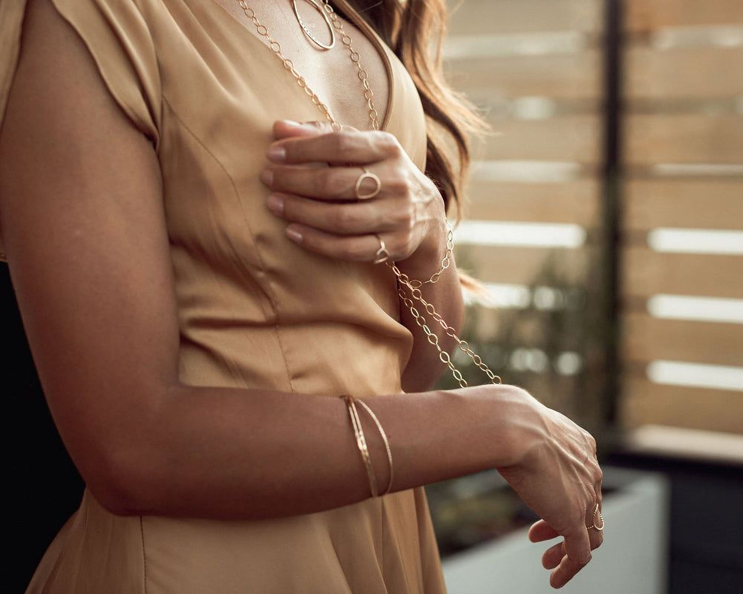 A woman wearing a gold dress and jewelry stands in front of a window.