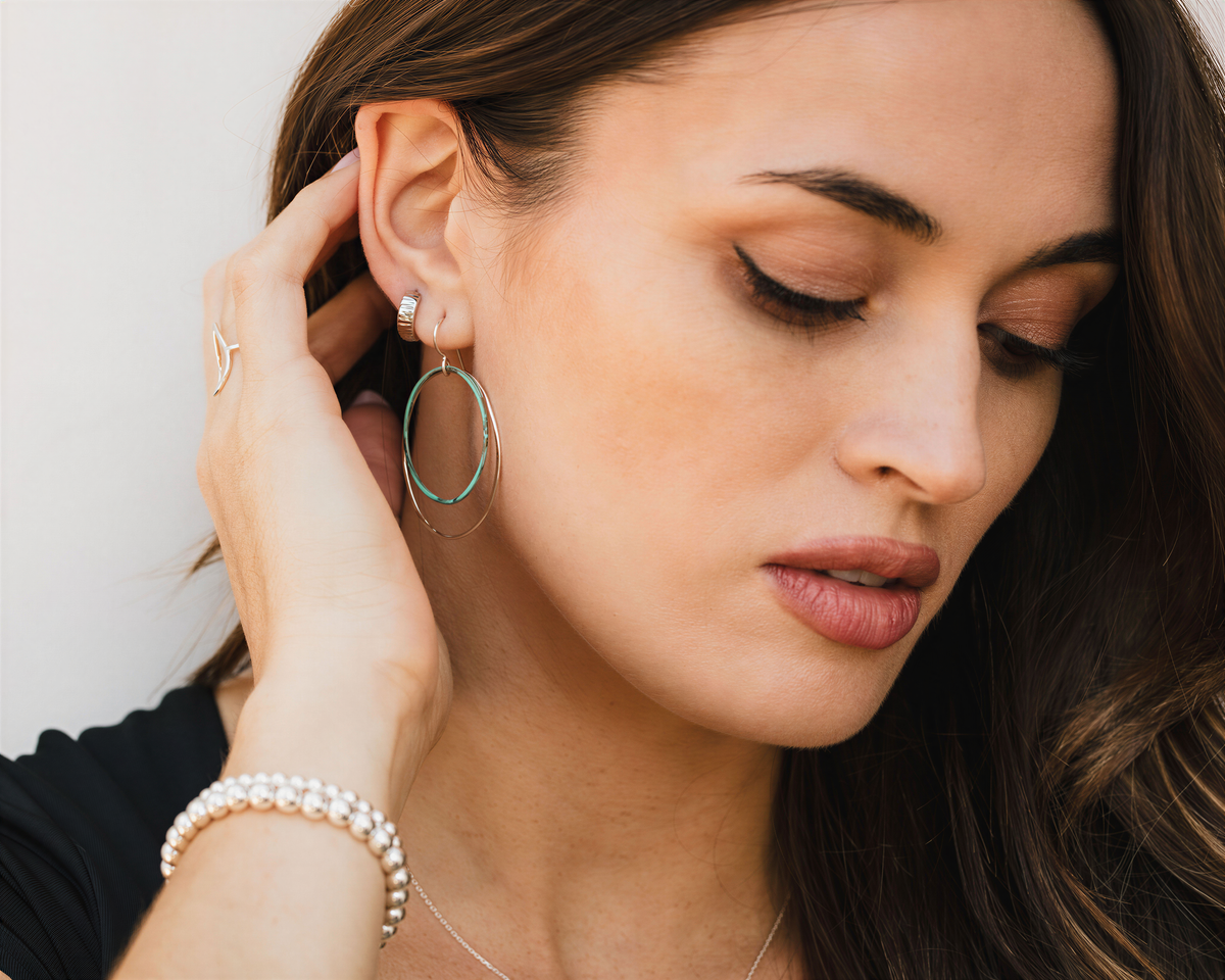 A woman with long brown hair is wearing a black top and has her hand on her ear, with her eyes closed and her lips slightly parted. She is also wearing a bracelet and earrings.