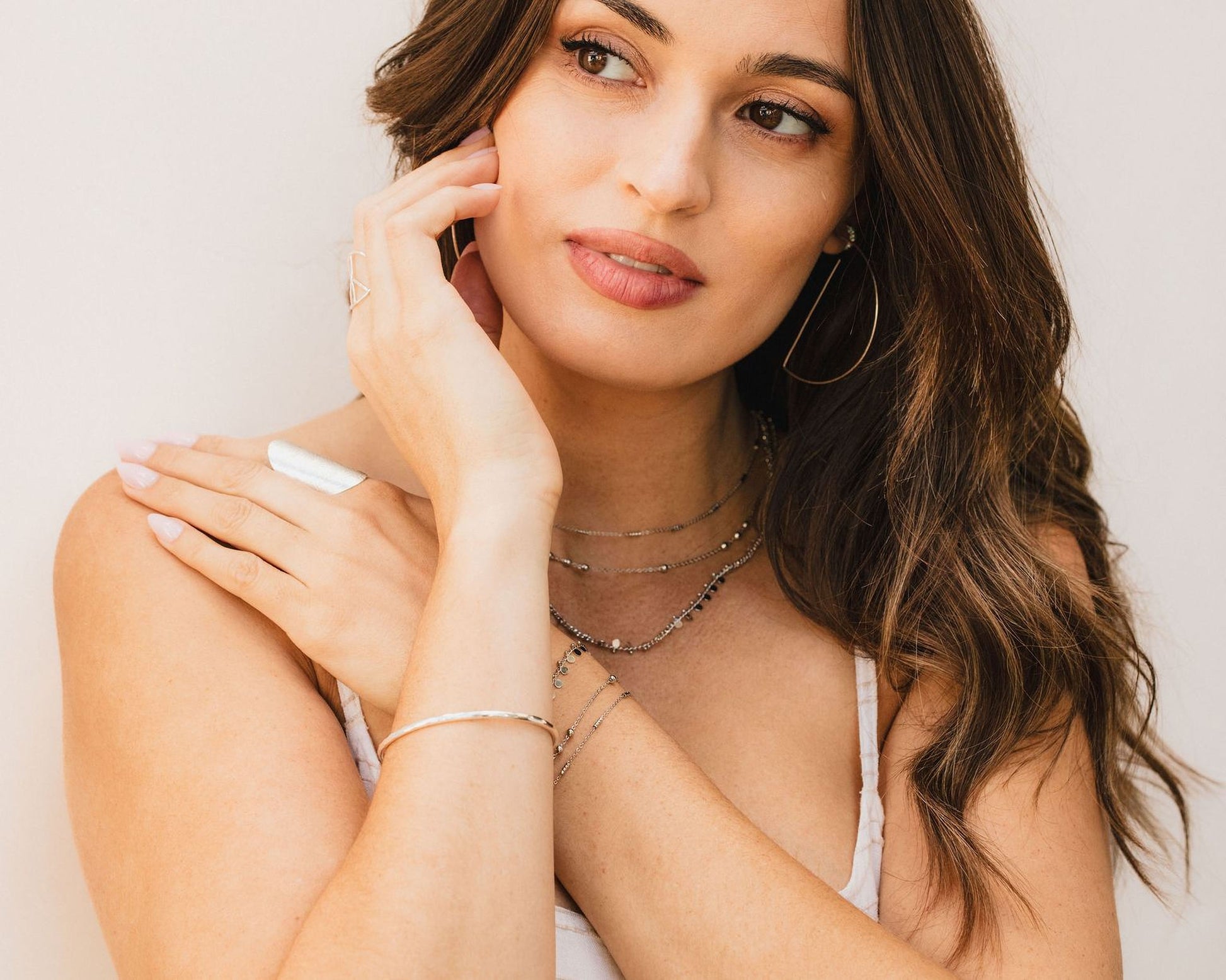 A woman with long brown hair wearing a white top and multiple necklaces, looking pensively to the side.