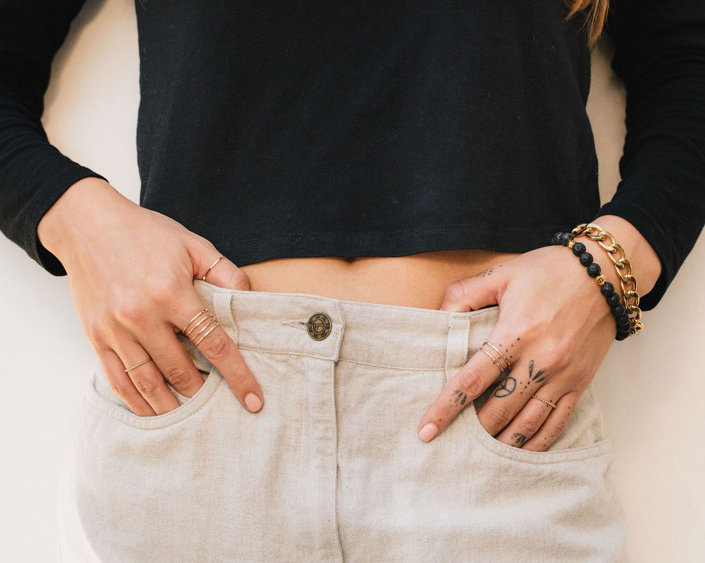 A person wearing a black shirt and beige pants, with multiple rings on their fingers and tattoos visible on their hands.