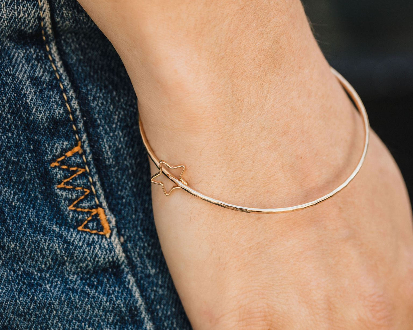 A gold-colored bangle bracelet is worn on a person's wrist, with a denim-colored jean pocket visible in the background.