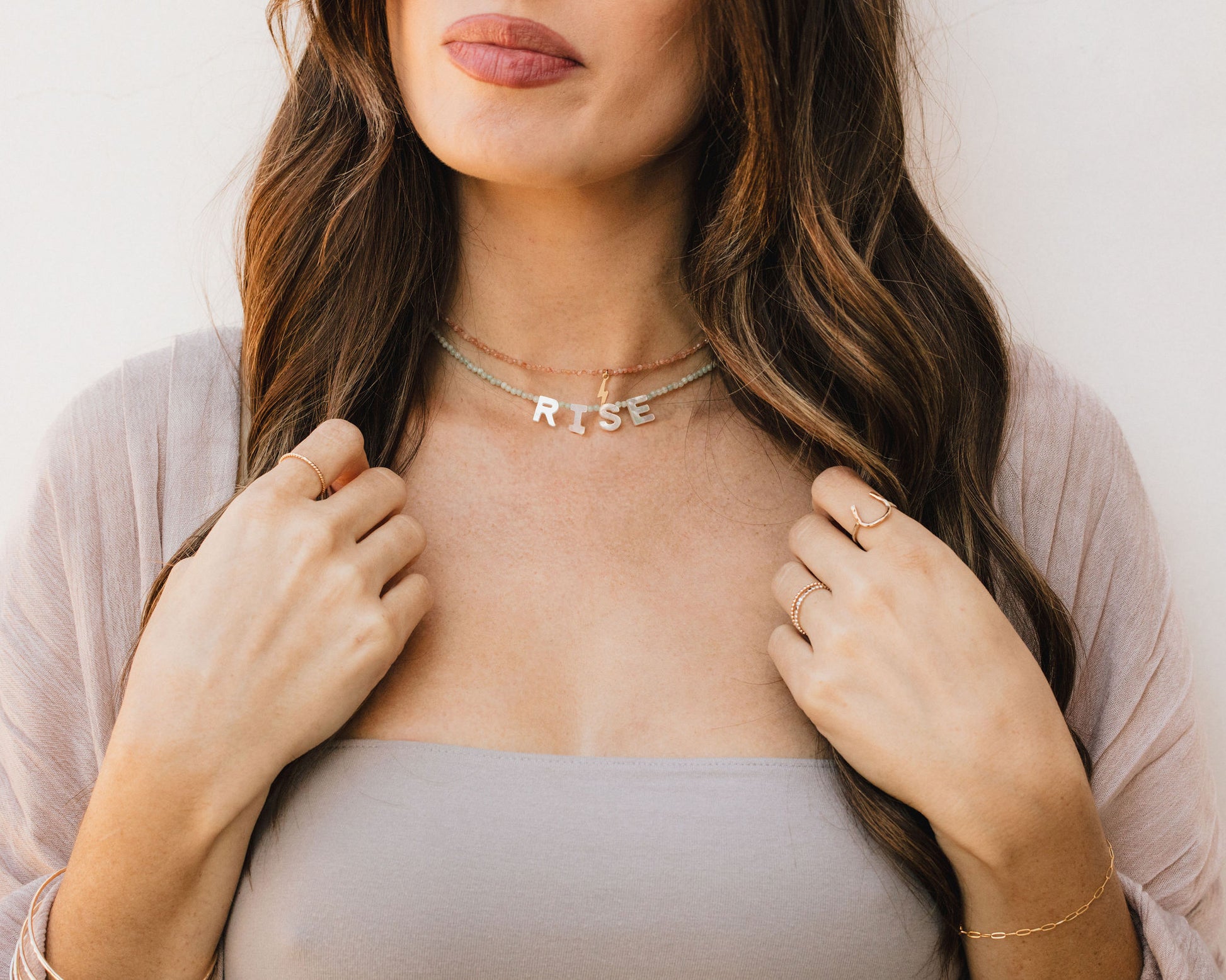 A woman with long brown hair wearing a necklace with the word "RISE" on it, posing with her hands on her chest.