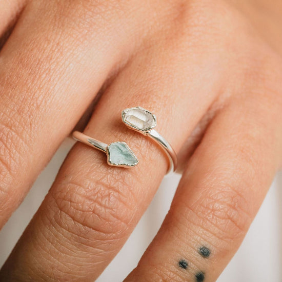 A close-up of a person's hand wearing a silver ring with a unique design, featuring a large, clear gemstone and a smaller, greenish-blue gemstone.
