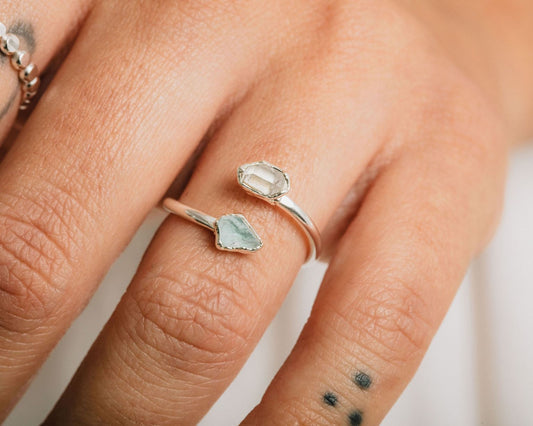 A close-up of a person's hand wearing a silver ring with a unique design, featuring a large, clear gemstone and a smaller, greenish-blue gemstone.