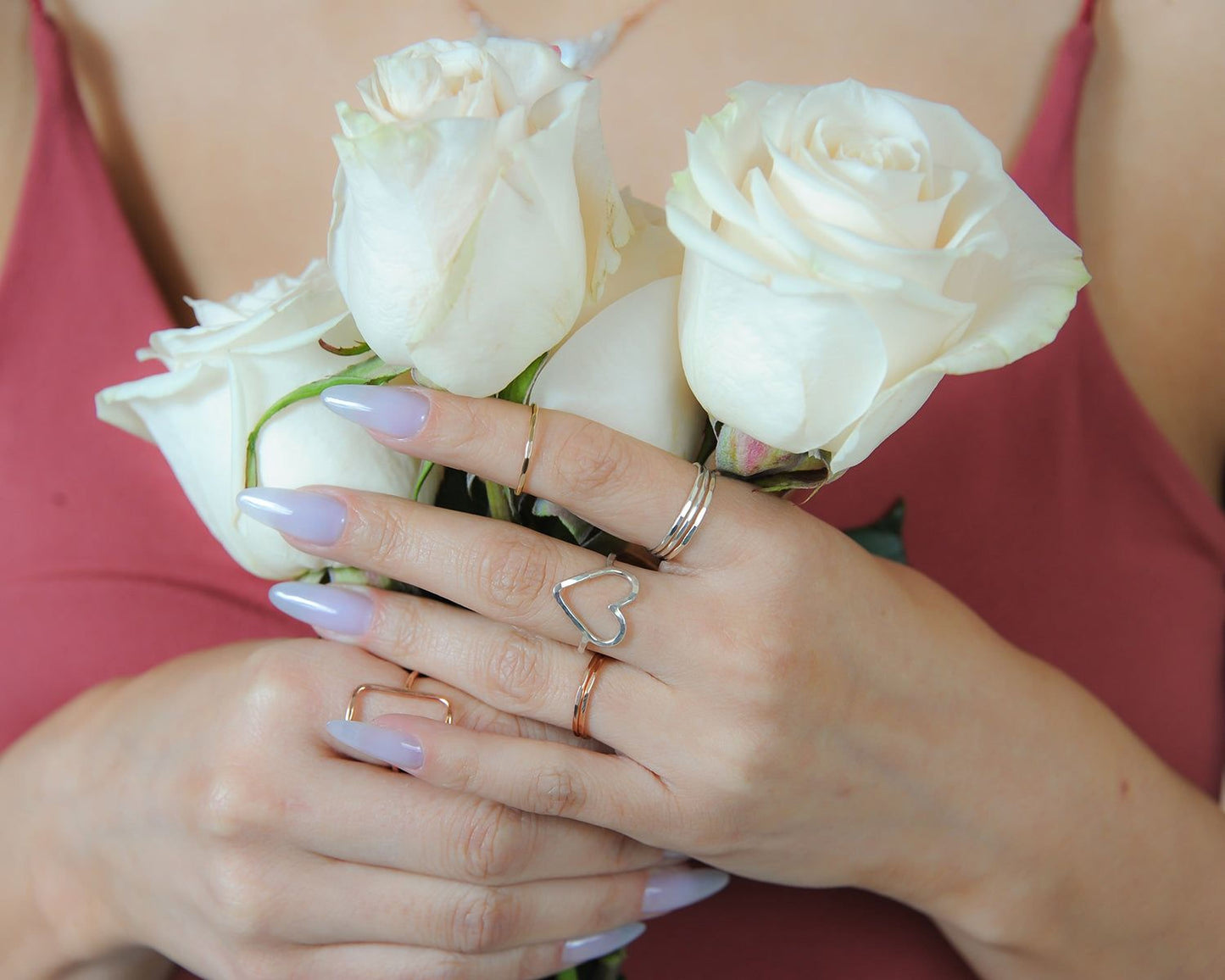 A woman wearing a red top holds a bouquet of white roses in her hands, with her fingers adorned with rings.
