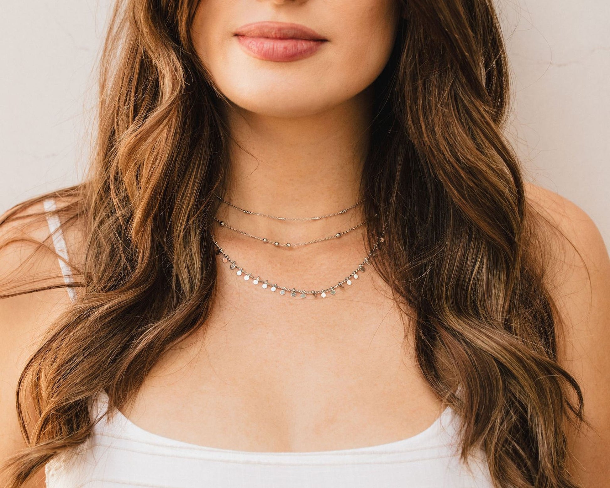 A close-up of a woman's face, focusing on her long, wavy brown hair and her white tank top. She is wearing a necklace with multiple strands of small, dangling charms.