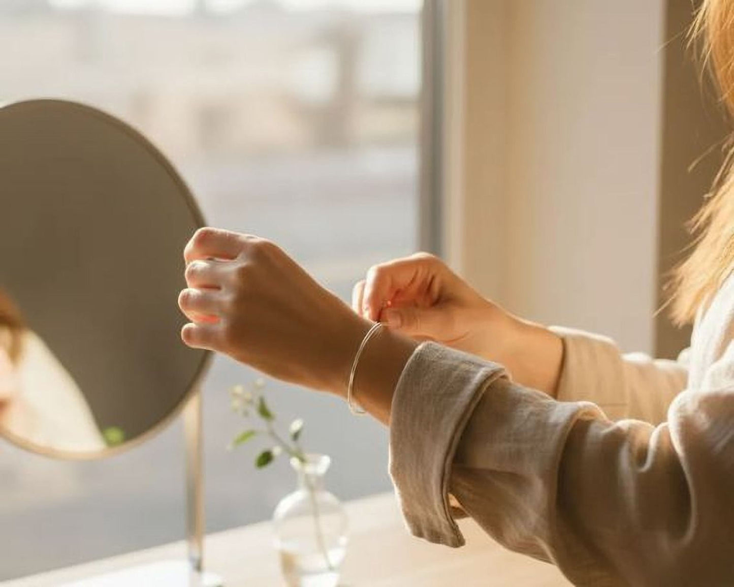A person's hands adjusting a bracelet on their wrist, with a round mirror and a small vase of flowers in the background.
