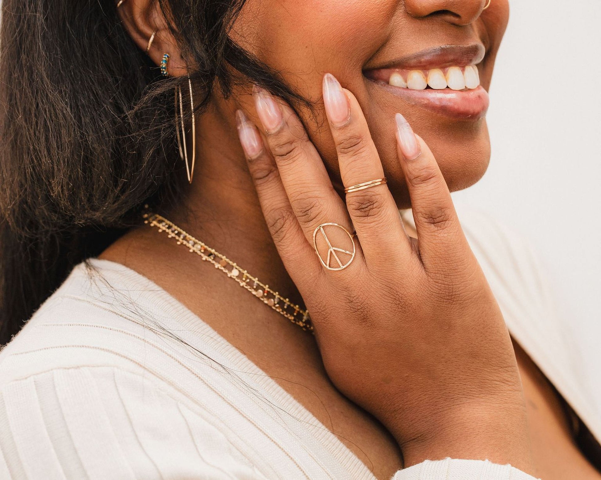 A close-up of a smiling woman's face, with her hand resting on her chin. She is wearing a white sweater and has several rings on her fingers, including a peace sign ring.