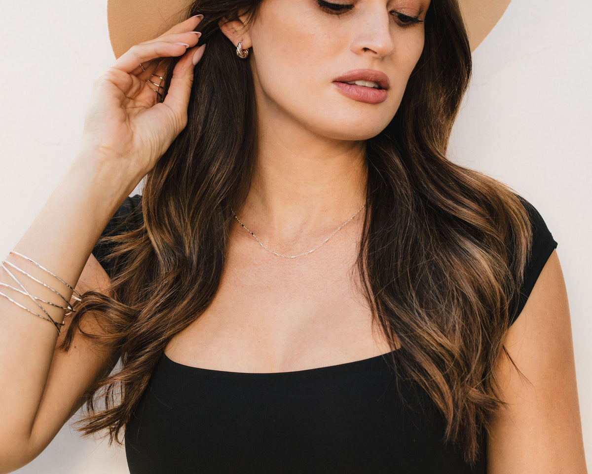 A woman with long, wavy brown hair wearing a black top and a wide-brimmed hat, with her hand touching her ear.