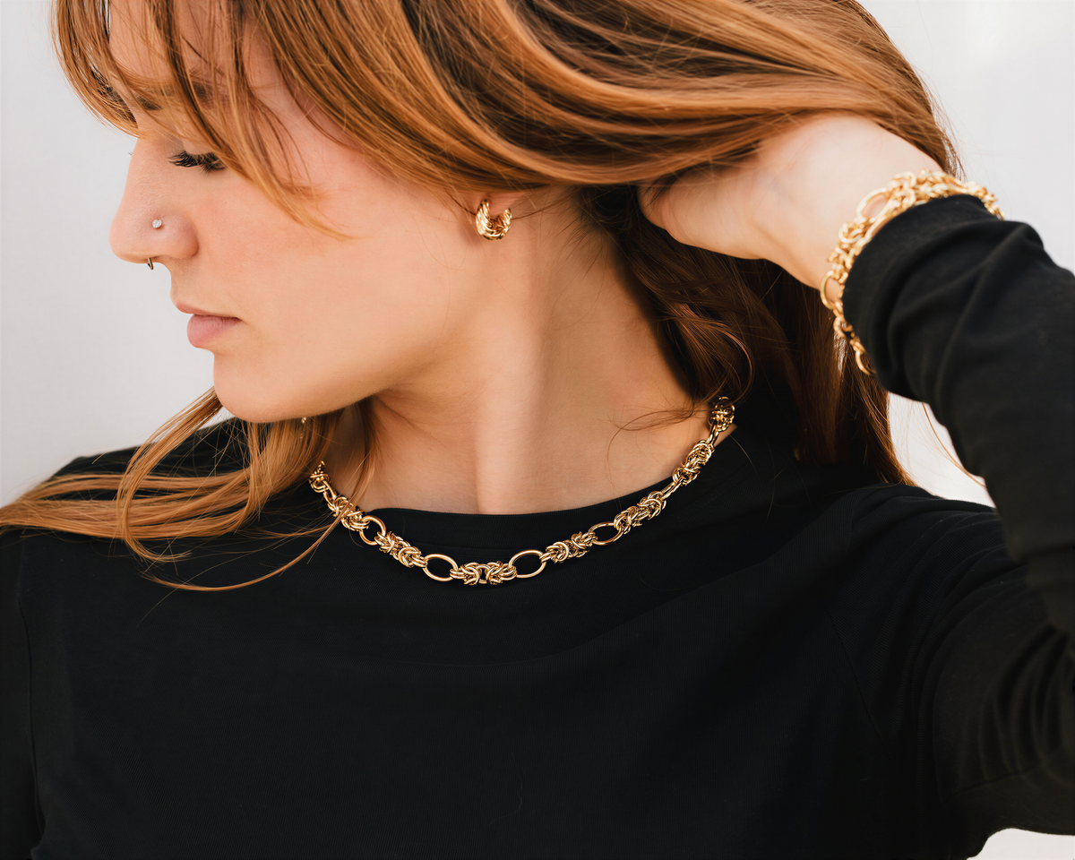 A woman with long, reddish-brown hair is wearing a black top and a gold necklace with a chain pendant. She is touching her hair with her hand.