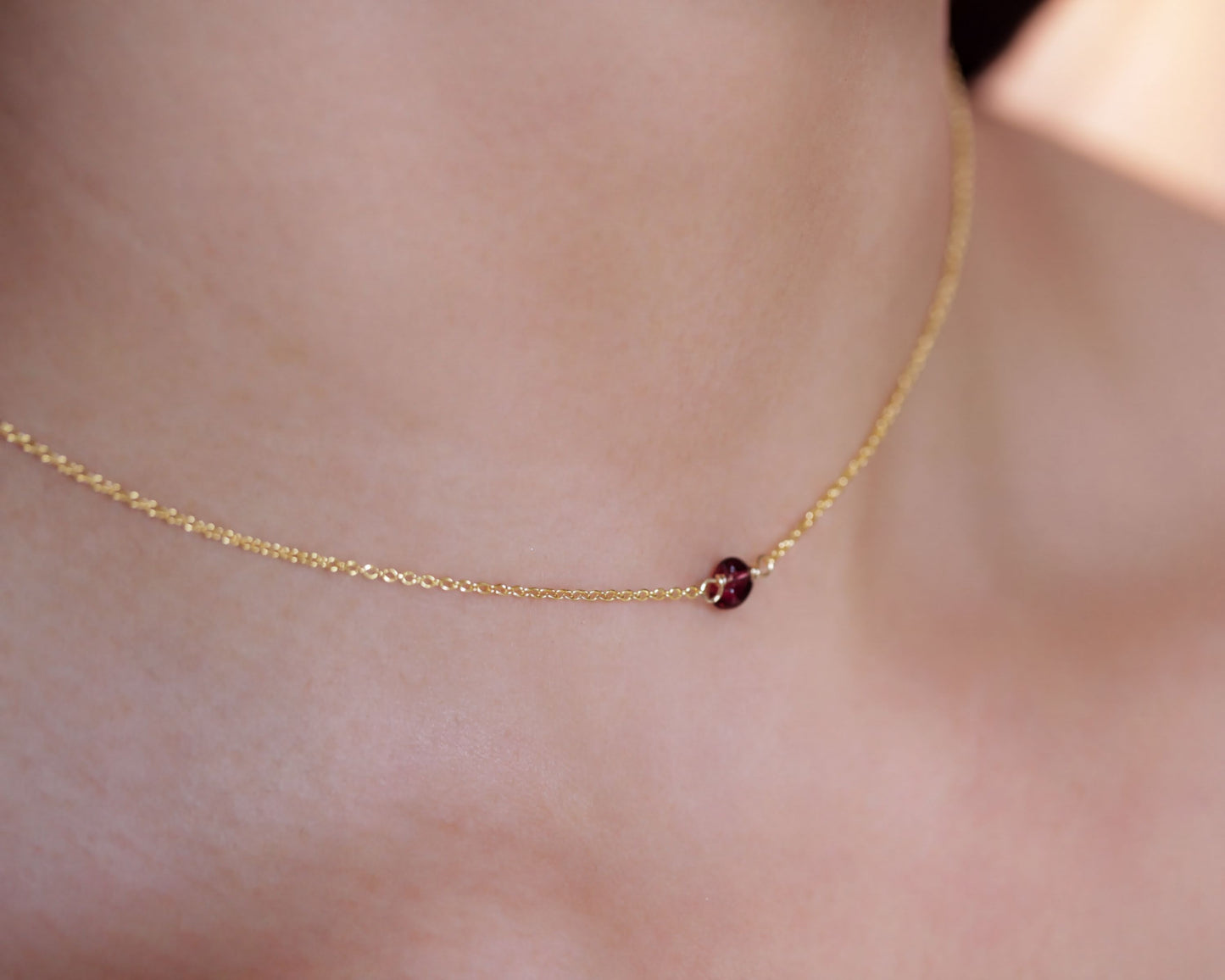 A delicate gold necklace with a small, round, red gemstone is shown in the image.