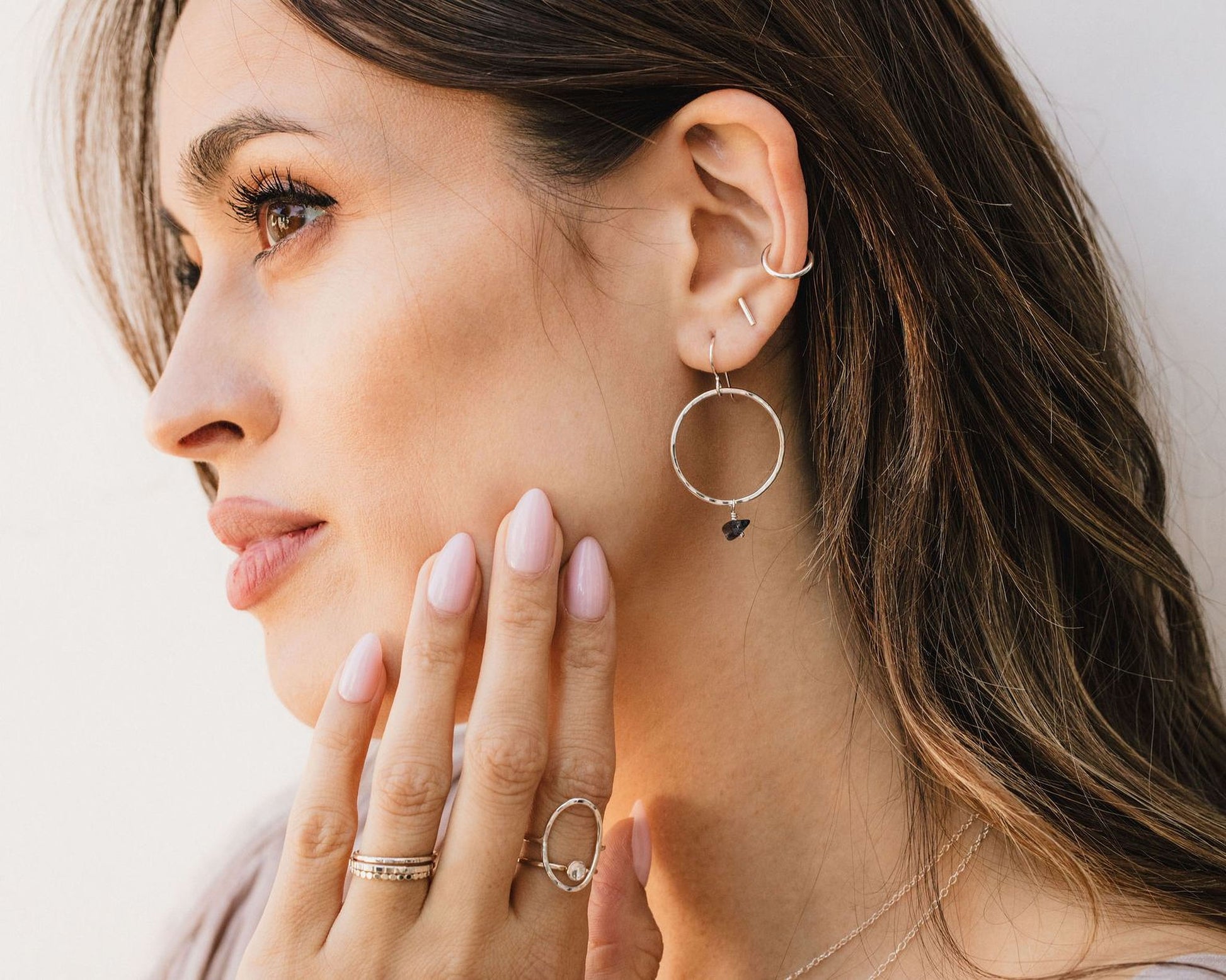 A woman with long brown hair wearing multiple rings and earrings, including a large hoop earring and a ring with a small gemstone.