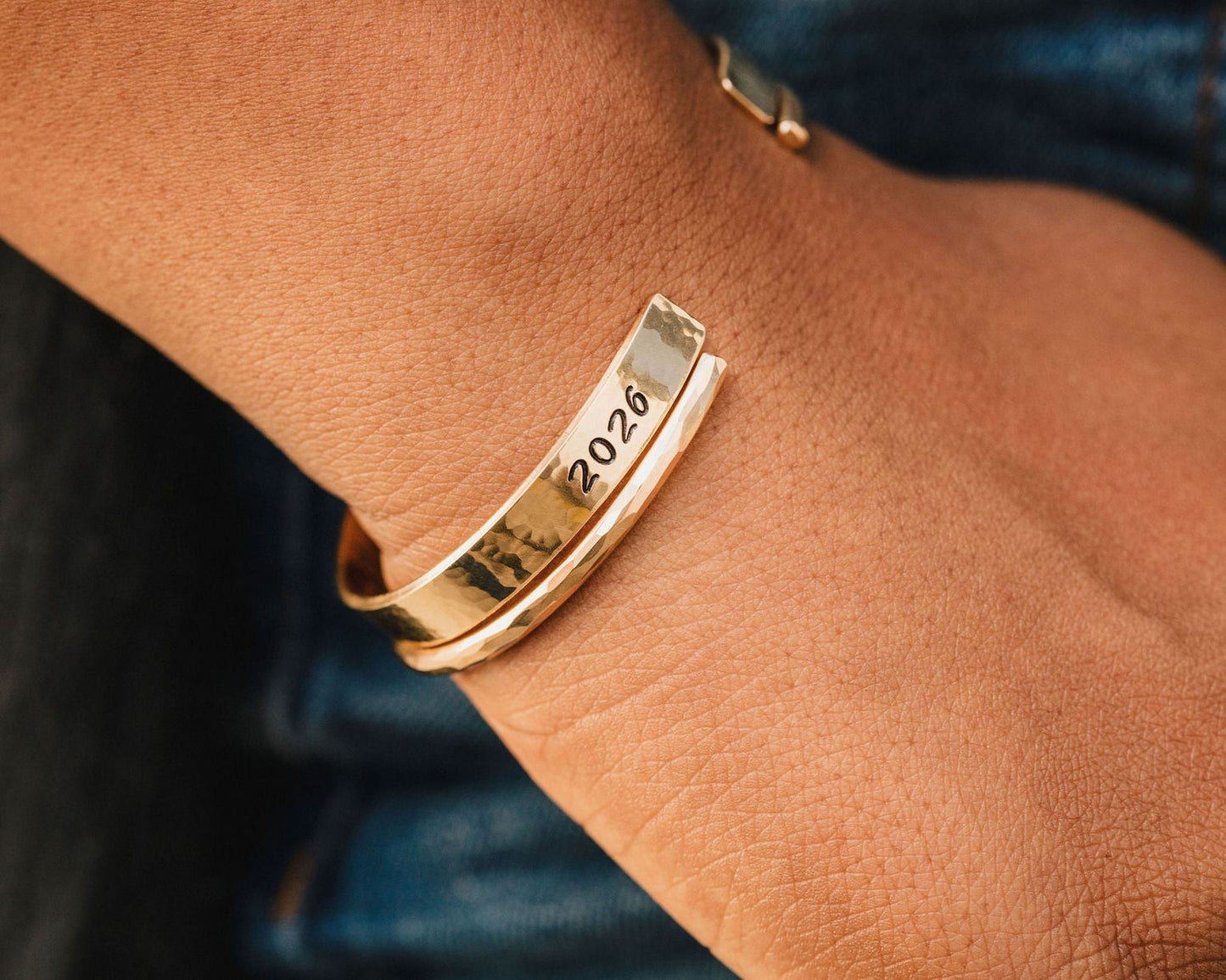 A gold bracelet with the year "2026" engraved on it is worn on a person's wrist.