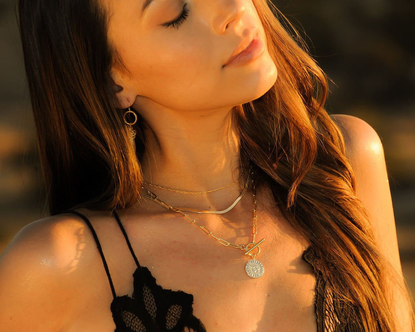 A woman with long, wavy hair wearing a black lace top and multiple necklaces, including a pendant necklace.