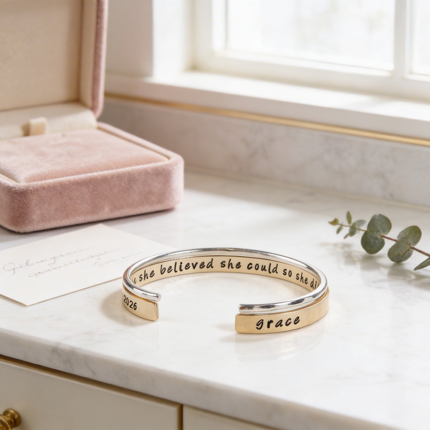A bracelet with the text "she believed she could so she was" engraved on it, placed on a marble countertop next to a pink jewelry box and a plant.