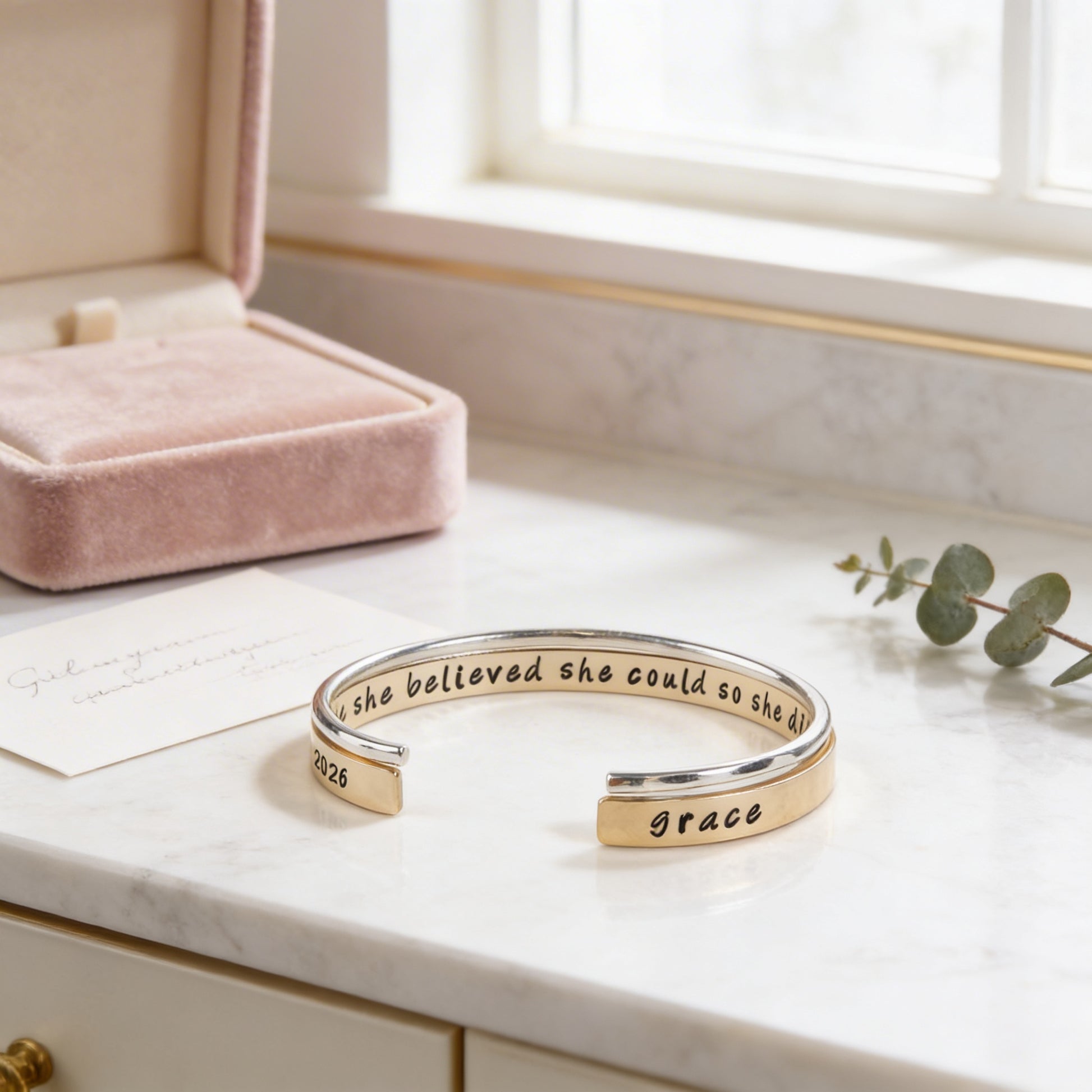 A bracelet with the text "she believed she could so she was" engraved on it, placed on a marble countertop next to a pink jewelry box and a plant.