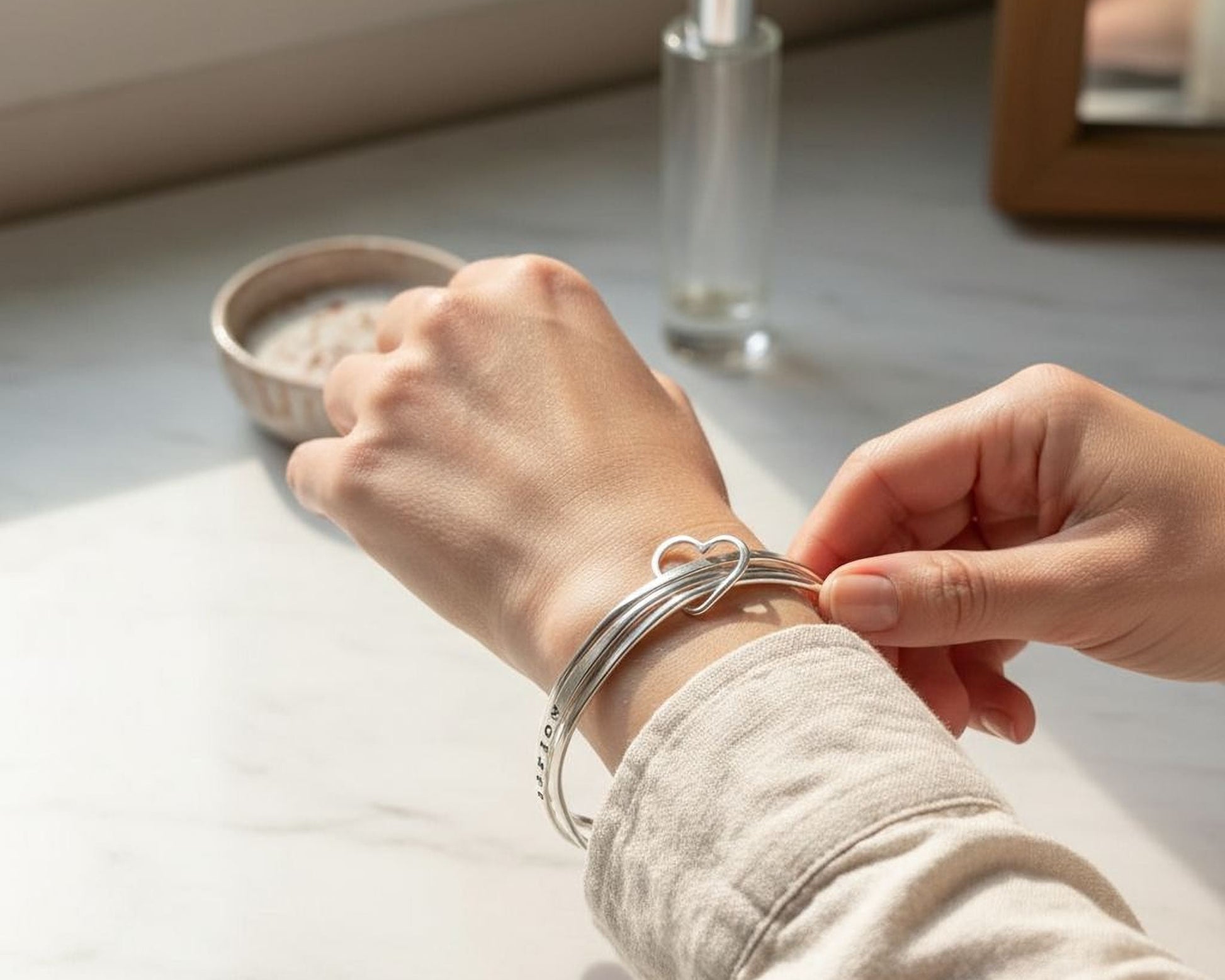A person's hand is adjusting a silver bracelet on their wrist, with a small bowl and a bottle of perfume visible in the background.