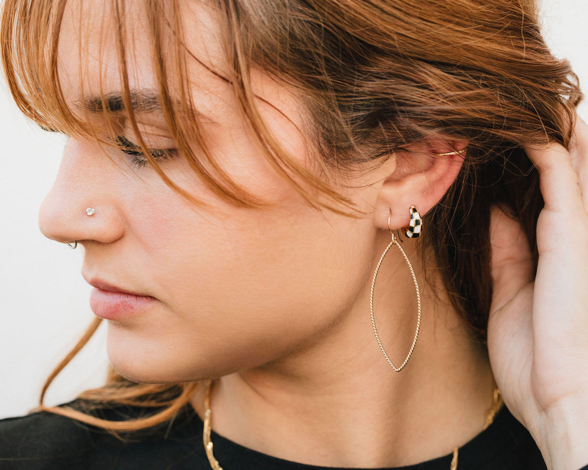 A close-up of a woman's face, with her hair styled in loose waves and her eyes closed. She is wearing large hoop earrings and a nose piercing.