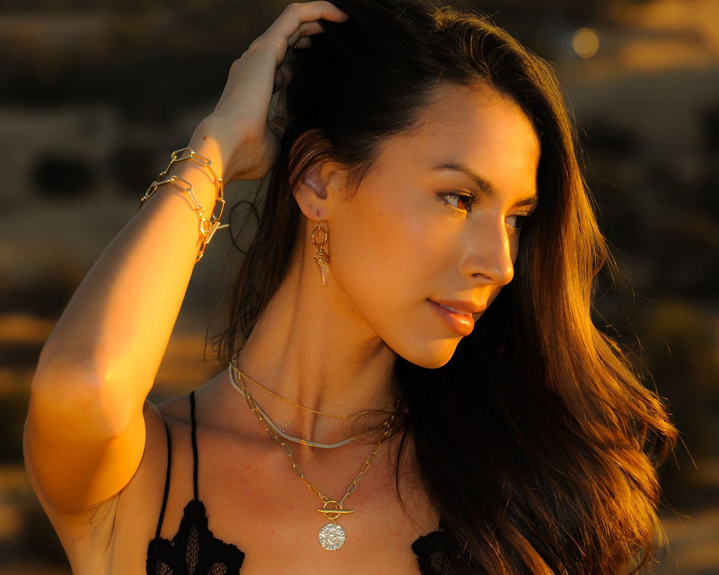 A woman with long dark hair wearing a black top and multiple necklaces poses with her hand on her head, her gaze directed off to the side.