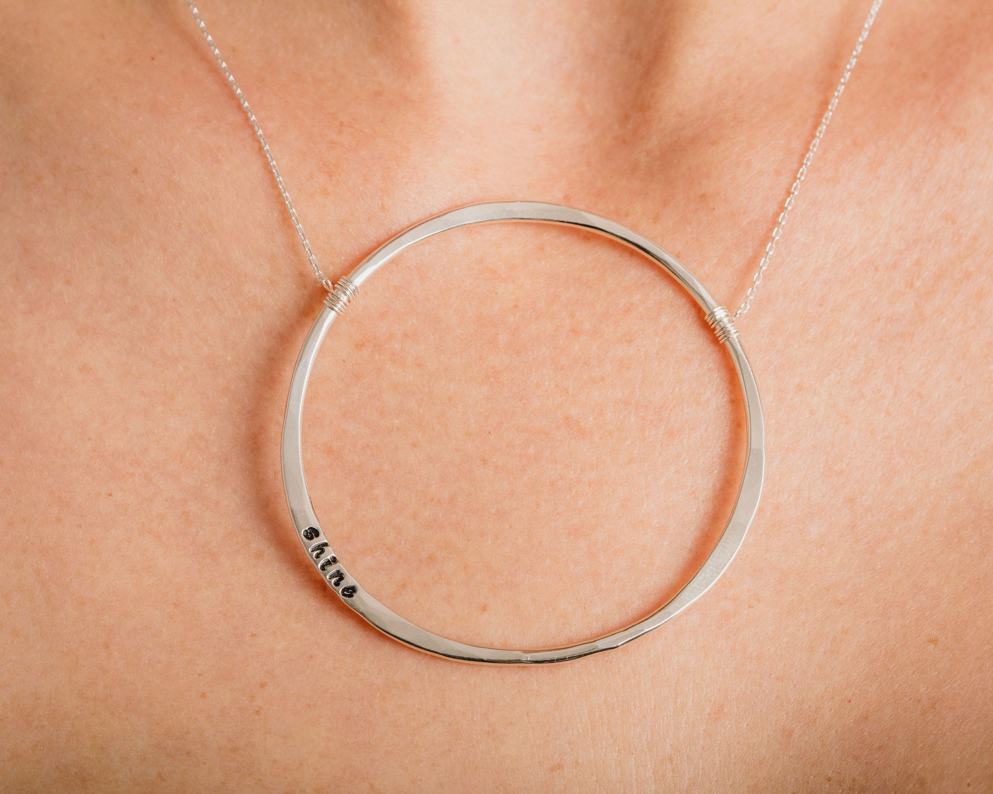 A close-up view of a person's neck and chest area wearing a silver circular necklace with a small engraved word on it.