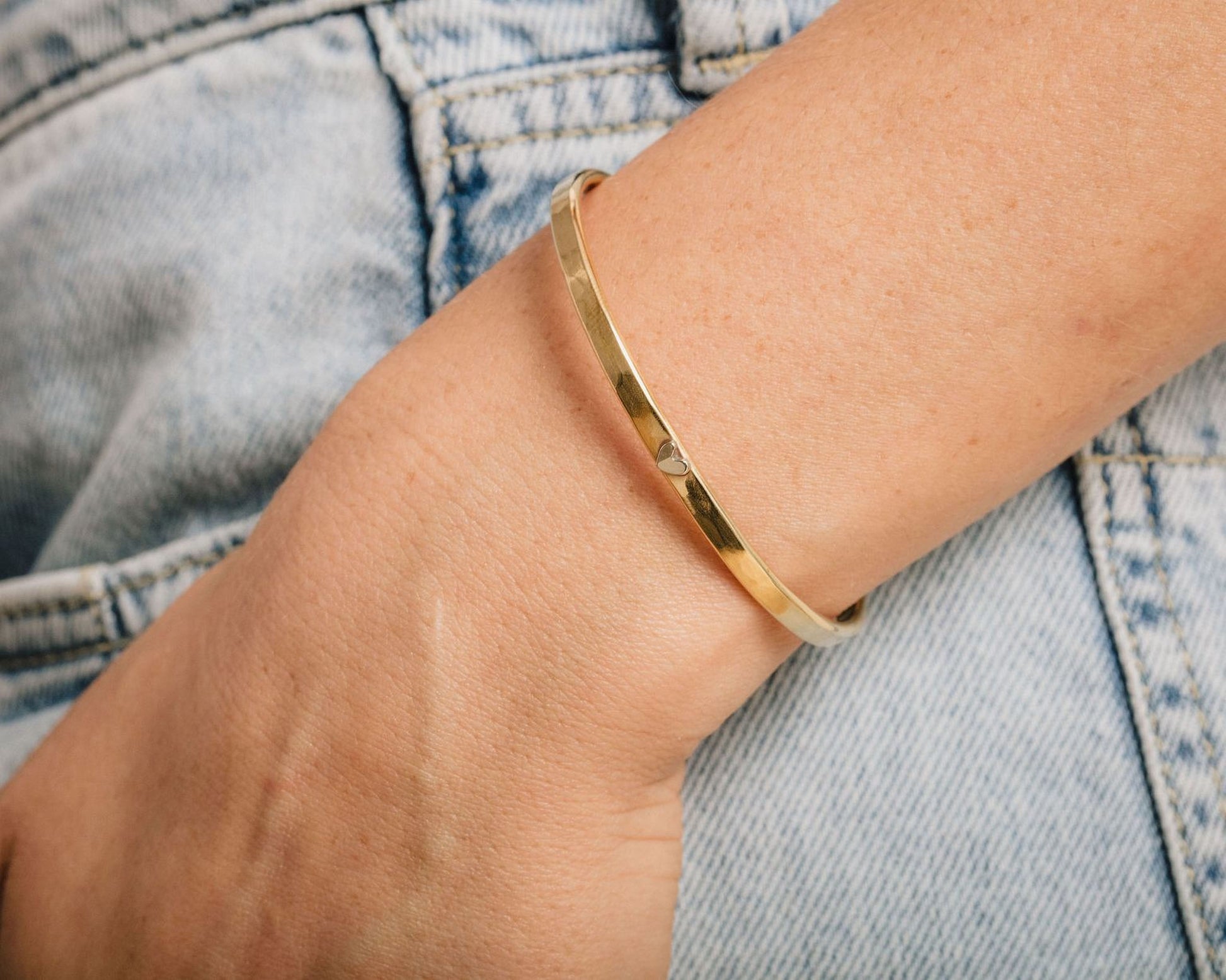 A gold bangle bracelet is worn on a person's wrist, with a denim jean pocket visible in the background.