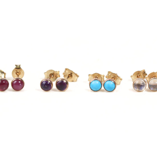 A collection of four gold-colored earrings with different colored gemstones, including red, purple, blue, and clear stones, arranged in a row.